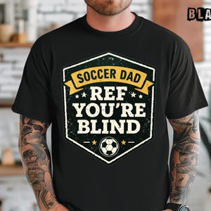 May include: Black t-shirt with a distressed graphic that reads "Soccer Dad Ref You're Blind." The design features a soccer ball and stars within a shield-shaped emblem. The word "BLACK" is printed in the upper right corner.