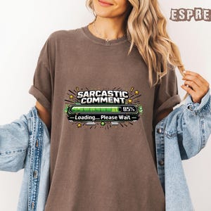 May include: Espresso-colored t-shirt with the text "Sarcastic Comment" above a loading bar graphic that reads "Loading... Please Wait" and shows 85% completion. The shirt is worn with a denim jacket.