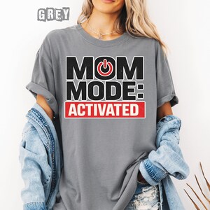 May include: A gray t-shirt with the text "MOM MODE: ACTIVATED" in black and red. The word "GREY" is printed at the top. The shirt is worn with a denim jacket.