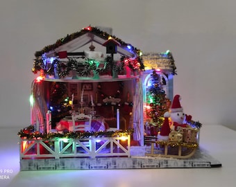 May include: A miniature Christmas village scene with a two-story house, decorated with colorful lights and garlands. A snowman in a Santa hat sits on a sleigh. The scene includes a Christmas tree and a dining table.