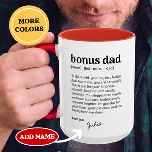 Op de afbeelding: Witte keramische mok met een rode binnenkant en handvat. De mok heeft de tekst "bonus dad" in het zwart, samen met een definitie en een oprechte boodschap. De mok heeft ook de tekst "ADD NAME" en "MORE COLORS".