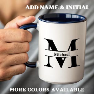 Op de afbeelding: Witte keramische mok met een marineblauwe binnenkant en handvat. De mok heeft een grote zwarte initiaal "M" met de naam "Michael" eronder. Tekst bovenaan luidt "ADD NAME & INITIAL" en onderaan "MORE COLORS AVAILABLE."