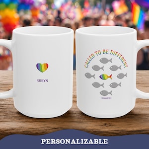 Op de afbeelding: Witte keramische mok met een regenbooghart en de naam "ROBYN" aan één kant. De andere kant heeft de tekst "CALLED TO BE DIFFERENT" met visillustraties, waarvan één in regenboogkleuren. De onderkant van de mok luidt "ROMANS 12:2".