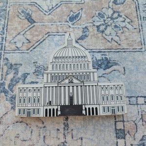 May include: A flat, white wooden cutout shaped like the United States Capitol building, featuring detailed black line art illustrations of the dome, columns, windows, and central entrance steps, creating a clear architectural representation.
