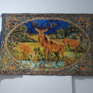 Könnte beinhalten: Rechteckige Tapisserie mit einer detaillierten Darstellung von Hirschen in einer natürlichen Landschaft. Ein großer Hirsch mit beeindruckendem Geweih steht im Mittelpunkt, während andere grasen und aus einem Bach trinken. Das Kunstwerk ist von einem verzierten Rahmen mit floralen und Schriftrollenmustern umgeben.