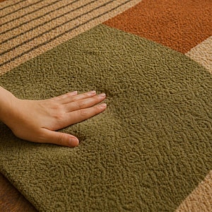 May include: Close-up of a textured rug with a geometric design. The rug features olive green, tan, and orange sections. A hand rests on the olive green section, highlighting the soft texture of the rug. The rug's design includes stripes and curved shapes.