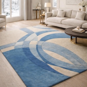 May include: A rectangular rug with a contemporary design. It features overlapping curved shapes in shades of blue and beige. The rug is placed on a wooden floor in a living room setting, with a sofa and coffee table.