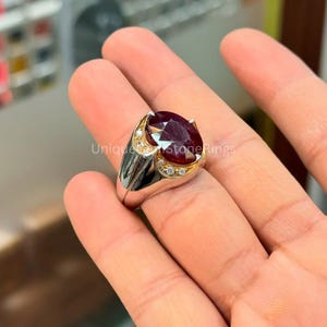 May include: A silver ring with a large, oval-cut, deep red gemstone. The ring has a gold-colored accent around the gemstone and small, clear stones. The ring is displayed on a hand.