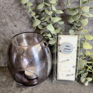 Op de afbeelding: Een zilverkleurige etherische oliebrander met een waxinelichtje erin, naast een verpakking witte patchouli wax melts. De wax melts hebben een gespikkeld ontwerp en zijn gelabeld met "White patchouli WAX MELT". Groene bladsprieten staan op de achtergrond.