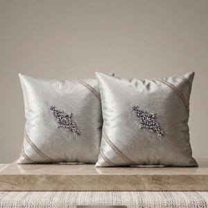 May include: Two square, pale silver cushions. Each cushion has a diagonal beaded trim and a central, embroidered design with purple and white accents. The cushions are placed on a marble surface.