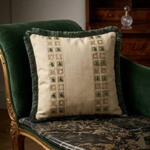 Op de afbeelding: Vierkant beige decoratief kussen met groene franjes. Het kussen heeft geborduurde geometrische patronen en is versierd met groene versieringen. Het kussen ligt op een donkergroene fluwelen chaise longue.