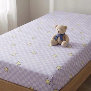 May include: A light purple and white checkered bed sheet featuring a repeating pattern of small yellow stars. A light brown plush teddy bear wearing a dark blue scarf is positioned in the center of the bed sheet on the mattress surface.