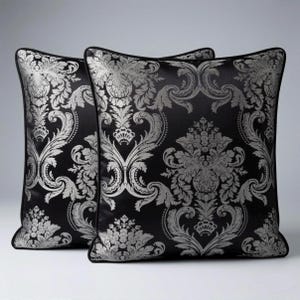Può includere: Due cuscini quadrati decorativi con sfondo nero e motivo damascato floreale argentato. I cuscini hanno un bordo nero e sono posizionati su una superficie bianca. Un articolo per l'arredamento della casa.