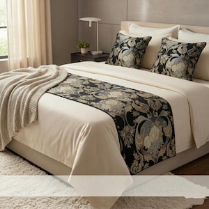 Könnte beinhalten: Ein Bett mit cremefarbener Bettwäsche, einem schwarz-goldenen Blumen-Bettläufer und zwei passenden Kissen. Eine beige Decke ist über die Seite des Bettes drapiert, und ein weißer Teppich liegt auf dem Boden.