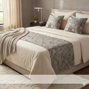 May include: A bed with cream-colored bedding, two decorative pillows, and a patterned bed runner. A beige knitted throw is draped over the foot of the bed. A small lamp and plant sit on a nightstand.