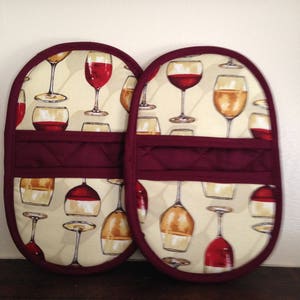 May include: Two red and cream oven mitts with a wine glass pattern. The mitts have a quilted design and a burgundy trim.