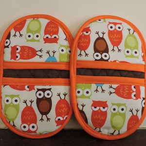 May include: A pair of orange and brown oven mitts with a white background featuring a repeating pattern of owls with different colored eyes and beaks.