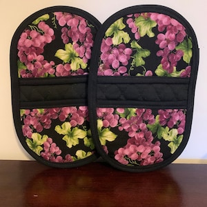 May include: A pair of black oven mitts with a grape pattern. The mitts have a quilted black band in the middle.