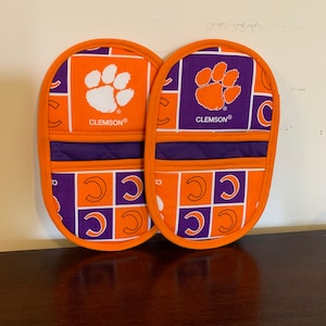 May include: A pair of orange and purple oven mitts with a Clemson Tigers paw print logo and the word "CLEMSON®" printed on each mitt.