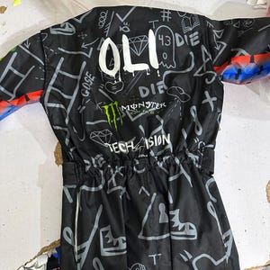 Puede incluir: Chaqueta negra con dise&ntilde;os de estilo grafiti en blanco y gris, incluyendo la palabra "OLI", un diamante y el logo de Monster Energy. La chaqueta tiene cintura con cord&oacute;n y detalles coloridos en las mangas.
