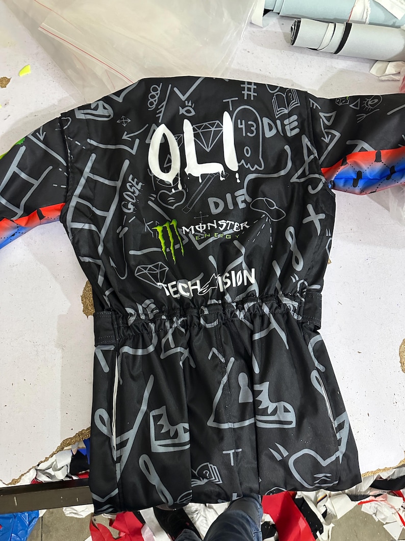 Puede incluir: Chaqueta negra con dise&ntilde;os de estilo grafiti en blanco y gris, incluyendo la palabra "OLI", un diamante y el logo de Monster Energy. La chaqueta tiene cintura con cord&oacute;n y detalles coloridos en las mangas.