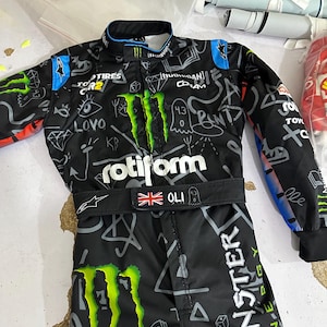 Puede incluir: Mono de carreras negro con el logotipo de Monster Energy y varios logotipos de patrocinadores. El mono presenta una bandera de la Union Jack y el nombre "OLI". El dise&ntilde;o es en blanco y negro con detalles en verde y azul.