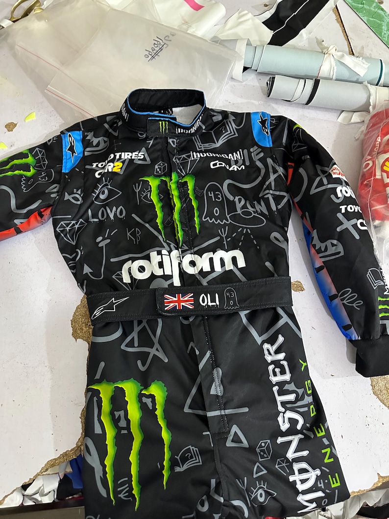 Puede incluir: Mono de carreras negro con el logotipo de Monster Energy y varios logotipos de patrocinadores. El mono presenta una bandera de la Union Jack y el nombre "OLI". El dise&ntilde;o es en blanco y negro con detalles en verde y azul.