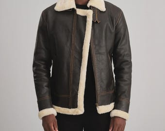 May include: A brown leather jacket with a cream-colored shearling collar and lining. The jacket features a zip closure, a buckled strap at the collar, and shearling trim on the cuffs. The jacket has a classic aviator style.