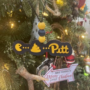 May include: A black and yellow Christmas tree ornament with the text "...💩...Pitt". The ornament is hanging on a Christmas tree with other ornaments and lights. The ornament is made of wood and has a black string.