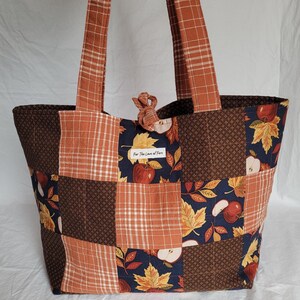 May include: A quilted tote bag featuring a patchwork design of orange plaid, dark brown dotted fabric, and a navy blue print with autumn leaves and red apples. The bag has two long shoulder straps and a small white label reading For The Love of Fern.