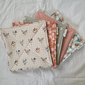 May include: A stack of quilted square pot holders with various patterns. One features a beige gingham with chicken illustrations. Others display floral, polka dot, and fruit designs in shades of peach, green, and pink. Each has a tag that reads "For The Love of Fun."