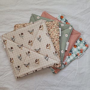 May include: A collection of quilted pot holders in various patterns. One features a beige gingham with chicken illustrations, while others display floral, polka dot, and fruit designs. The pot holders are in shades of beige, pink, green, and orange.