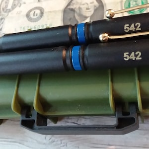 Two thin Blue line bullet twist pens with gold components and gold engraving on top of a miniature replica tactical gun case in green