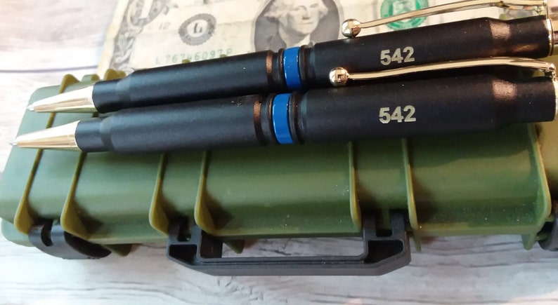 Two thin Blue line bullet twist pens with gold components and gold engraving on top of a miniature replica tactical gun case in green