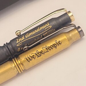 以下が含まれることがあります： 刻まれた文字が書かれた金と黒のペン2本。 上のペンには「2nd Amendment A well regulated Militia」という文字が刻まれ、下のペンには「We the People」という文字が刻まれています。