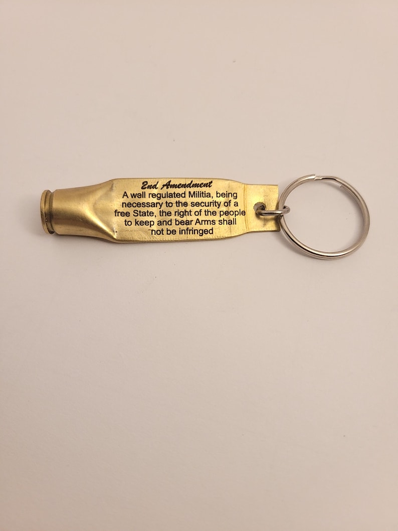 K&ouml;nnte beinhalten: Ein goldfarbener Schl&uuml;sselanh&auml;nger in Form einer Patronenh&uuml;lse mit dem Text "2nd Amendment" und dem Text "A well regulated Militia, being necessary to the security of a free State, the right of the people to keep and bear Arms shall not be infringed" eingraviert.