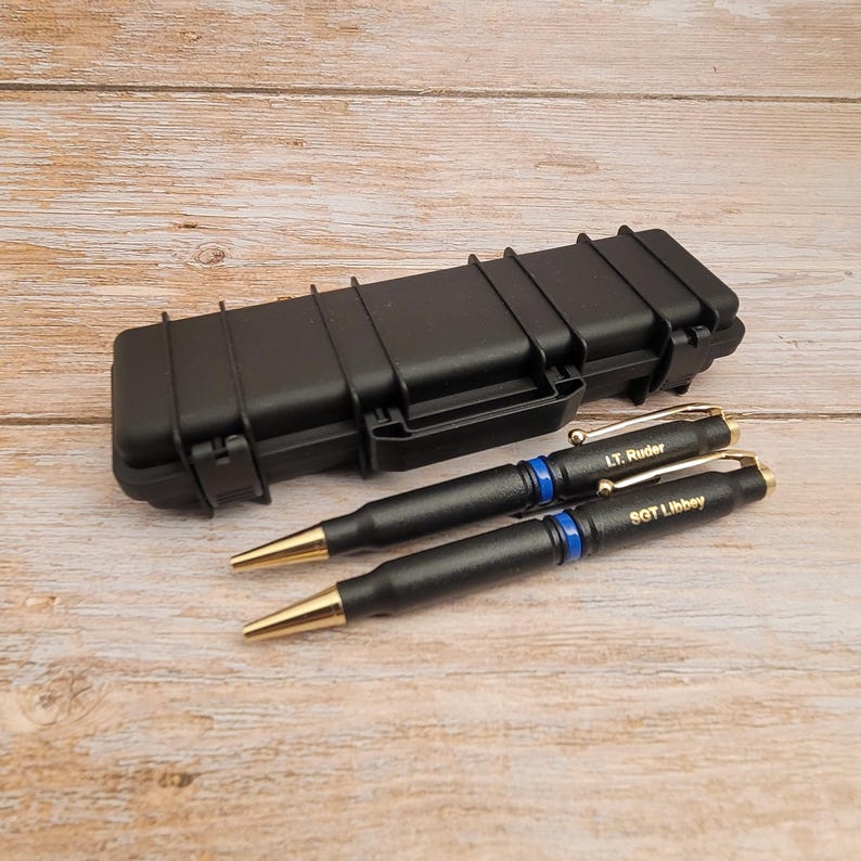 May include: Two black bullet pen style pens with gold tips and blue accents. The pens are engraved with the names "LT. Ruder" and "SGT. Libbey". The pens are in a black hard case.