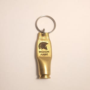 K&ouml;nnte beinhalten: Ein goldfarbener Schl&uuml;sselanh&auml;nger in Form einer Patronenh&uuml;lse mit einem spartanischen Helm und dem eingravierten Text "MOLON LABE".