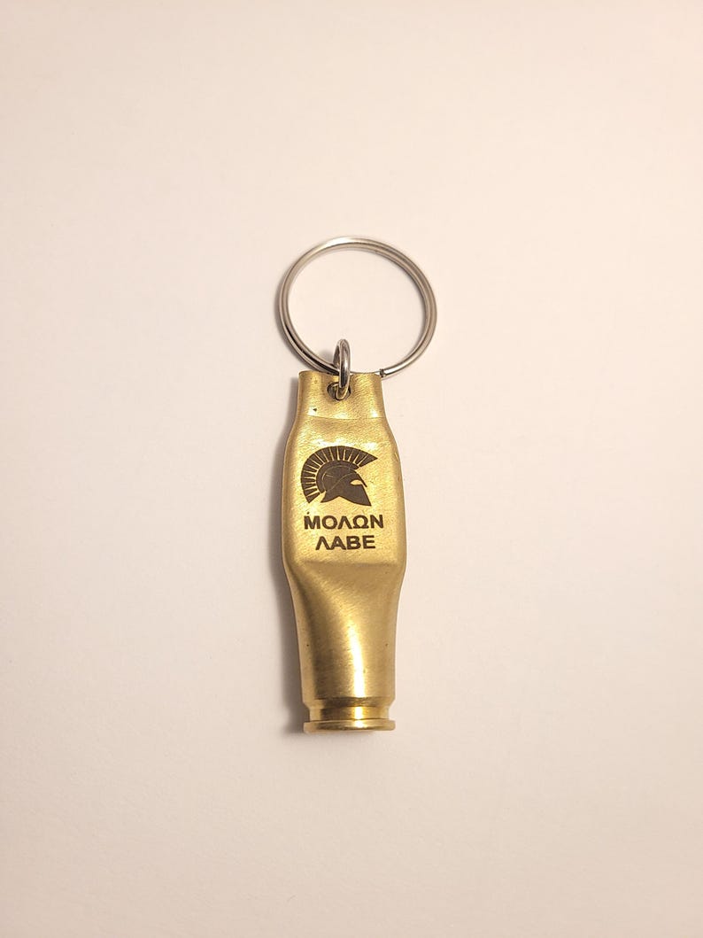 K&ouml;nnte beinhalten: Ein goldfarbener Schl&uuml;sselanh&auml;nger in Form einer Patronenh&uuml;lse mit einem spartanischen Helm und dem eingravierten Text "MOLON LABE".