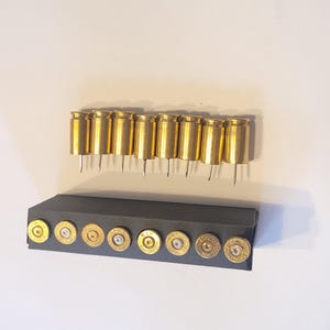 May include: A display of brass bullet casings, arranged in two rows. The top row features casings with attached pins, while the bottom row shows the base of the casings in a black holder. The casings have a shiny, metallic finish.
