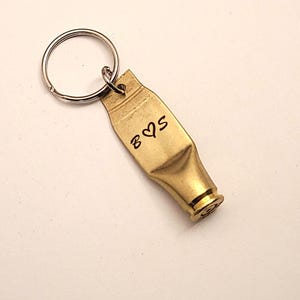 Puede incluir: Un llavero de color dorado hecho de una vaina de bala con un llavero de plata. La vaina tiene las iniciales "B & S" grabadas con un s&iacute;mbolo de coraz&oacute;n entre las letras.