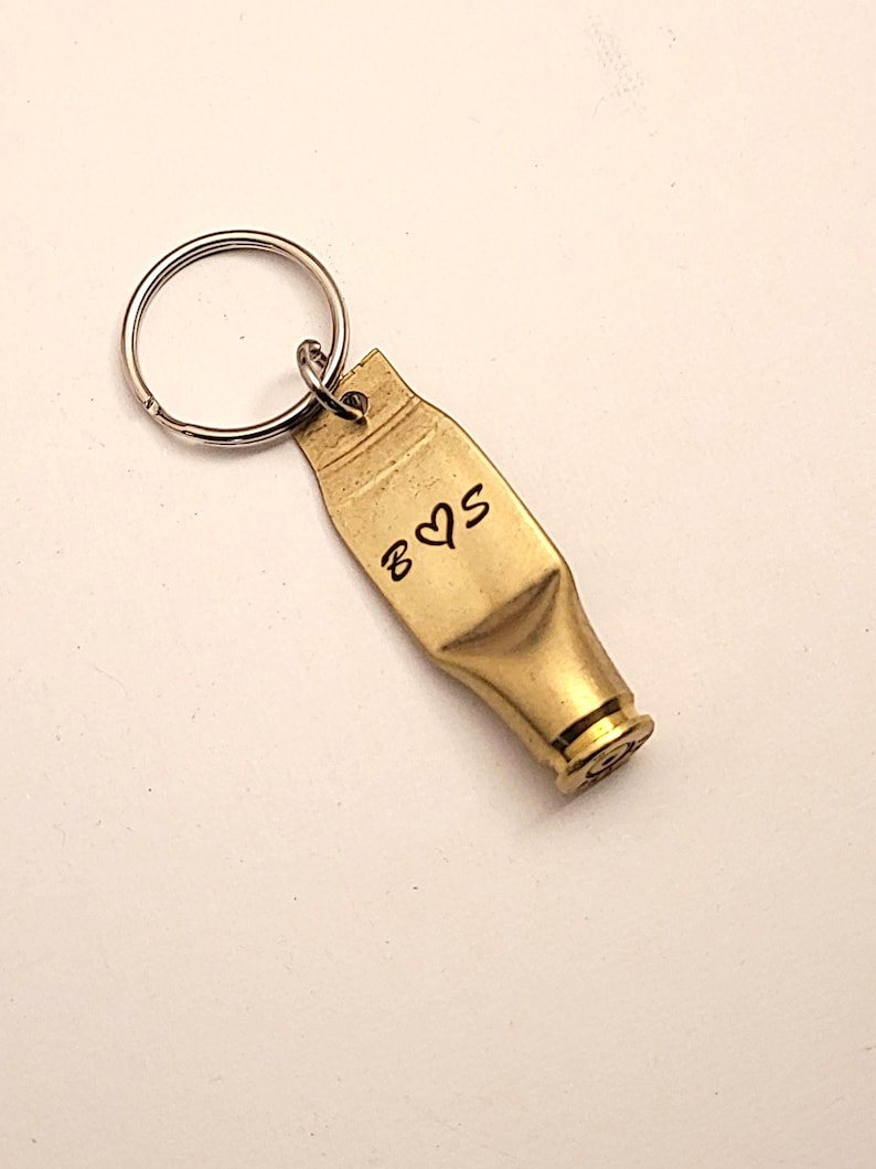 Puede incluir: Un llavero de color dorado hecho de una vaina de bala con un llavero de plata. La vaina tiene las iniciales "B & S" grabadas con un s&iacute;mbolo de coraz&oacute;n entre las letras.