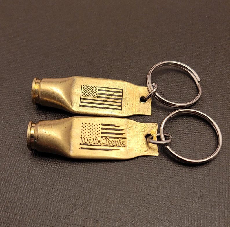 K&ouml;nnte beinhalten: Zwei Schl&uuml;sselanh&auml;nger aus Messing mit einem US-Flaggen-Design. Ein Schl&uuml;sselanh&auml;nger tr&auml;gt die Worte "We the People" eingraviert.