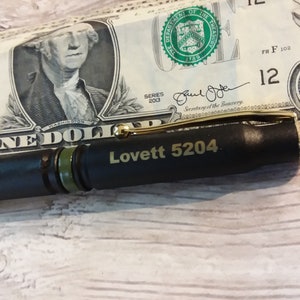 Pu&ograve; includere: Una penna nera con una fascia verde e dettagli dorati. La penna ha inciso il testo "Lovett 5204". La penna &egrave; appoggiata su una banconota da un dollaro.