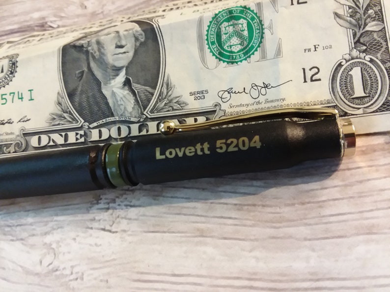 Pu&ograve; includere: Una penna nera con una fascia verde e dettagli dorati. La penna ha inciso il testo "Lovett 5204". La penna &egrave; appoggiata su una banconota da un dollaro.