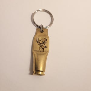 K&ouml;nnte beinhalten: Ein goldfarbener Schl&uuml;sselanh&auml;nger mit einem gravierten Hirschkopf. Der Schl&uuml;sselanh&auml;nger ist aus einer verbrauchten Patronenh&uuml;lse gefertigt.
