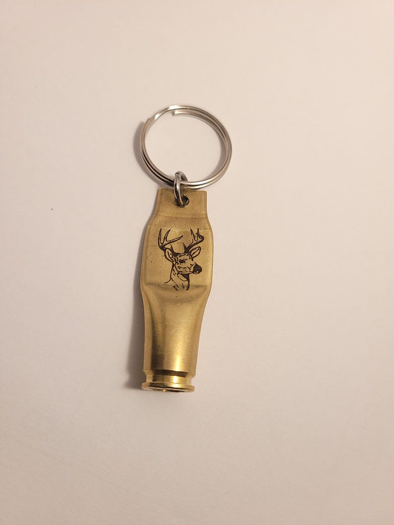 K&ouml;nnte beinhalten: Ein goldfarbener Schl&uuml;sselanh&auml;nger mit einem gravierten Hirschkopf. Der Schl&uuml;sselanh&auml;nger ist aus einer verbrauchten Patronenh&uuml;lse gefertigt.