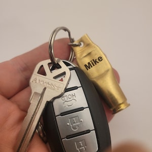 Puede incluir: Un llavero dorado con un dije de casquillo de bala grabado con el nombre "Mike". El llavero est&aacute; unido a una llave plateada y a un llavero de coche negro con botones etiquetados como "Hold" y "Unlock".