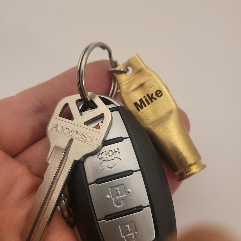 Puede incluir: Un llavero dorado con un dije de casquillo de bala grabado con el nombre "Mike". El llavero est&aacute; unido a una llave plateada y a un llavero de coche negro con botones etiquetados como "Hold" y "Unlock".