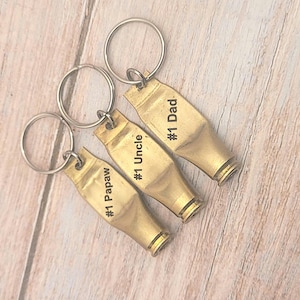 Puede incluir: Tres llaveros de color dorado con texto grabado. Los llaveros tienen forma de casquillos de bala y tienen el texto "#1 Papaw", "#1 Uncle" y "#1 Dad" grabado en ellos.